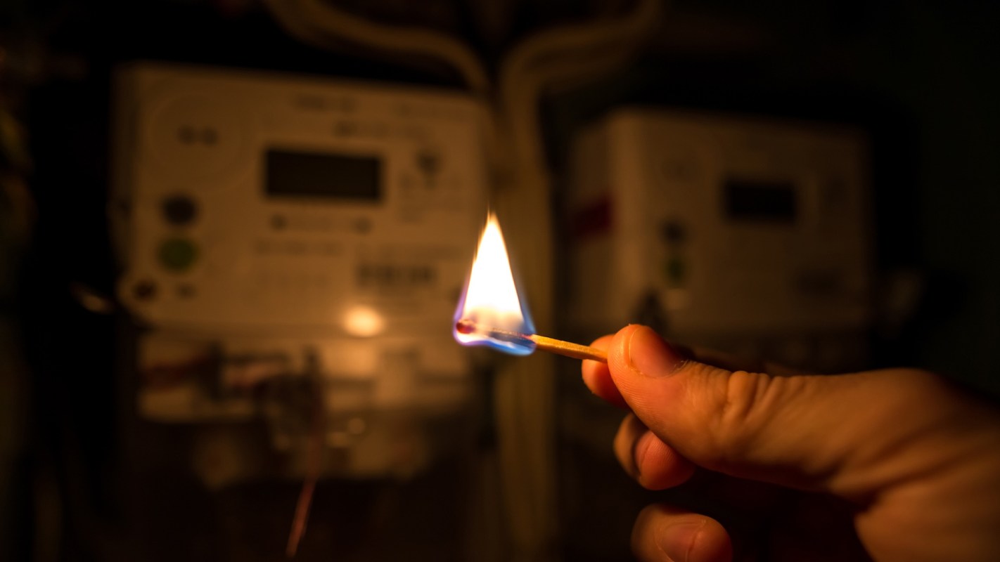 A lit match is held in front of electrical meters in a dimly lit environment.