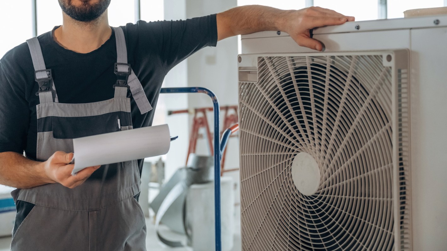 A technician holds paperwork next to a heat pump unit in a construction setting.