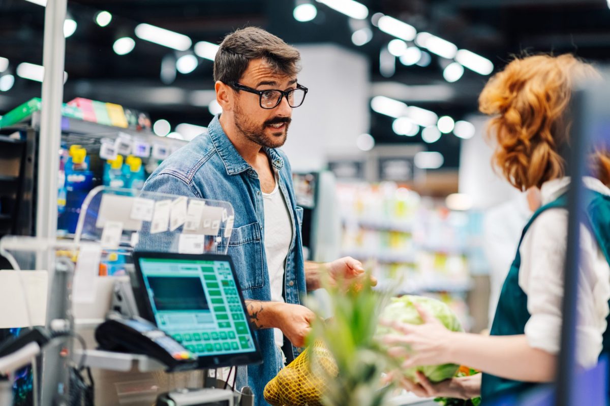 A man interacts with a cashier at a grocery store while holding a bag of produce.