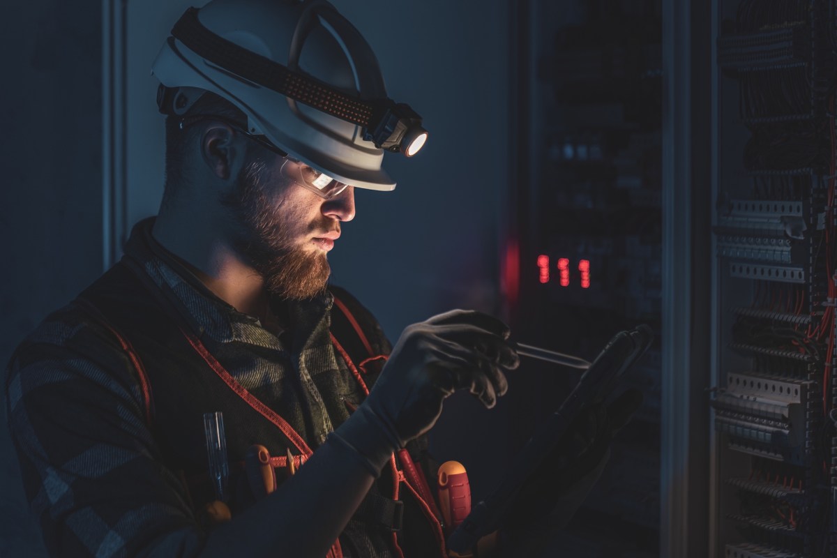 A worker in a safety helmet focuses on a tablet near electrical equipment during a power outage.