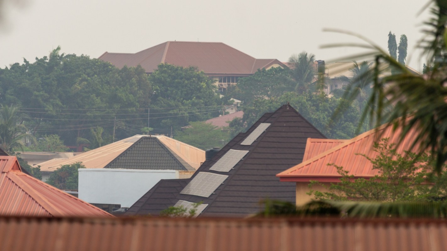 A view of rooftops, including some with solar panels, with varying colors and trees in the background.