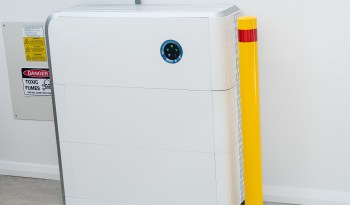 A white solar battery storage unit with warning signs and a yellow safety post nearby.