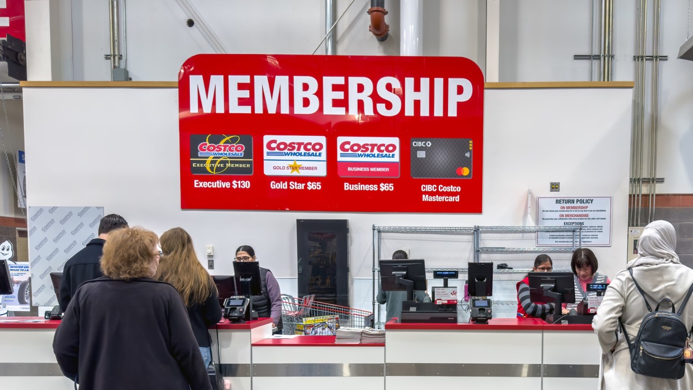A Costco membership sign displays options and prices while customers engage with staff at the membership counter.
