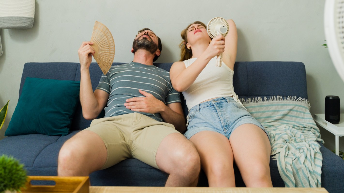 A man and a woman on a couch, using fans to cool themselves in a warm indoor setting.