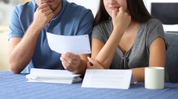 A man and a woman express doubt while examining a document while seated at a table with papers and a cup.