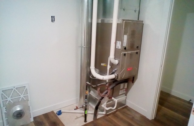 A heating unit with pipes is installed in a corner of a room with light-colored walls and wooden flooring.
