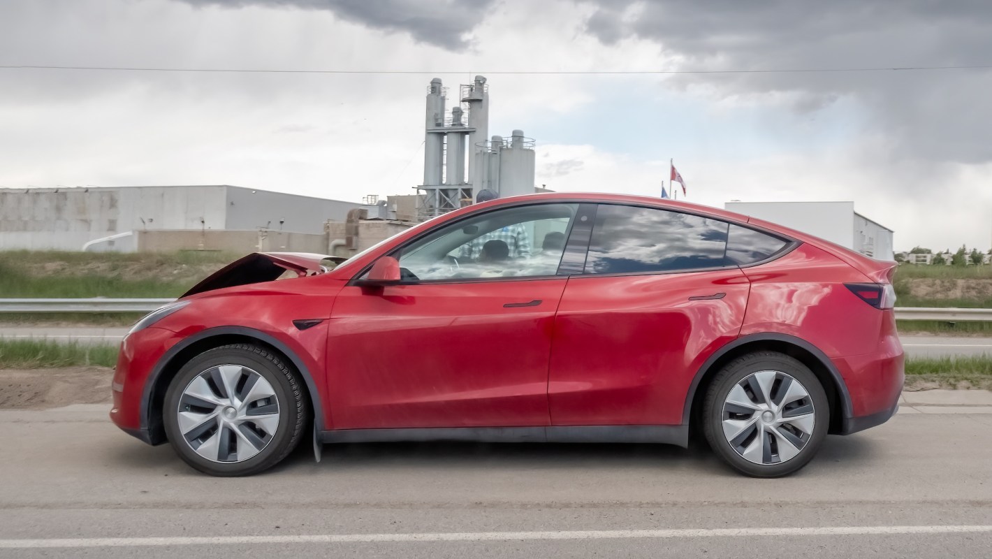 A red Tesla Model Y with front damage parked beside a road and industrial buildings in the background.