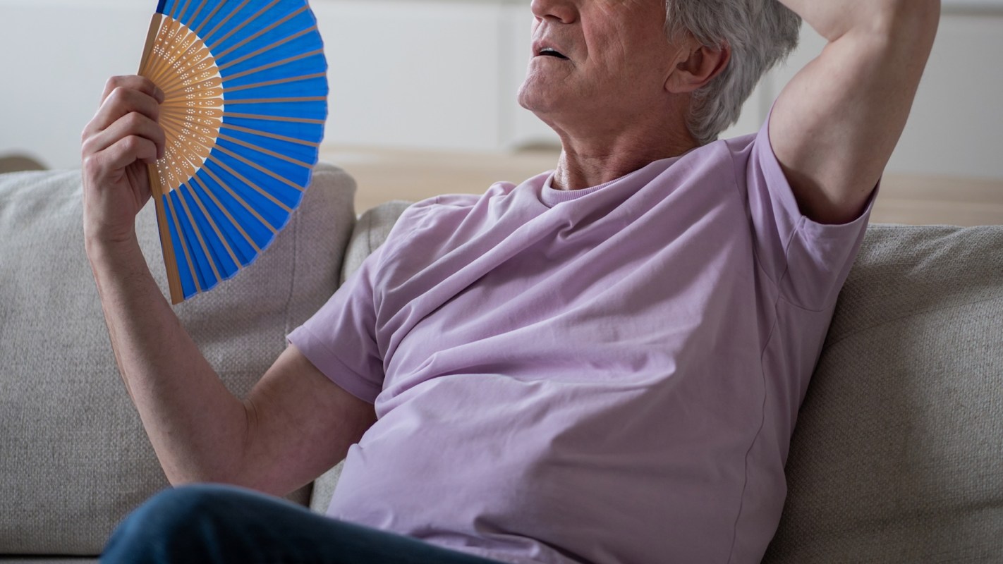 A man sitting on a couch holds a blue fan, looking upward with his head tilted back.