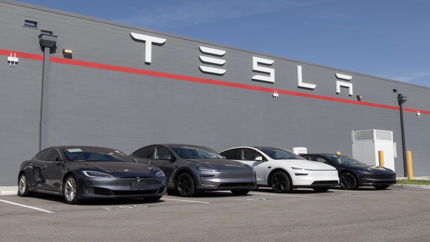 Four Tesla vehicles parked in front of a gray building with a large "TESLA" sign.