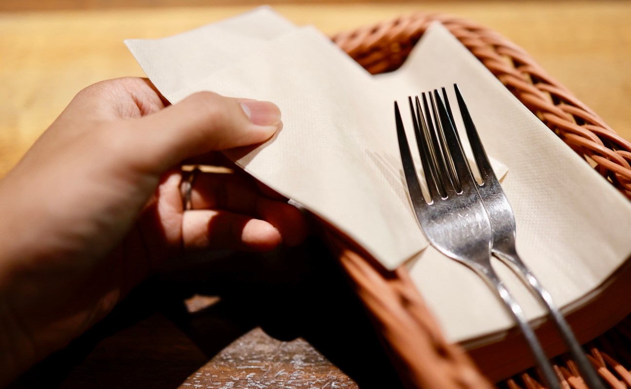 A hand holding a napkin over a basket containing two forks and a stack of napkins.