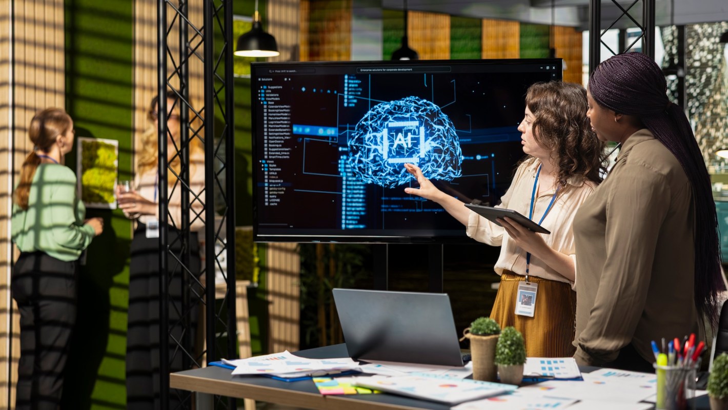 Two women discuss a digital presentation about AI on a screen while standing by a table with documents in an office setting.