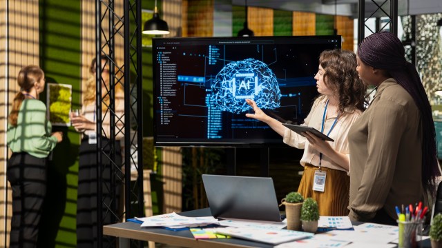 Two women discuss a digital presentation about AI on a screen while standing by a table with documents in an office setting.