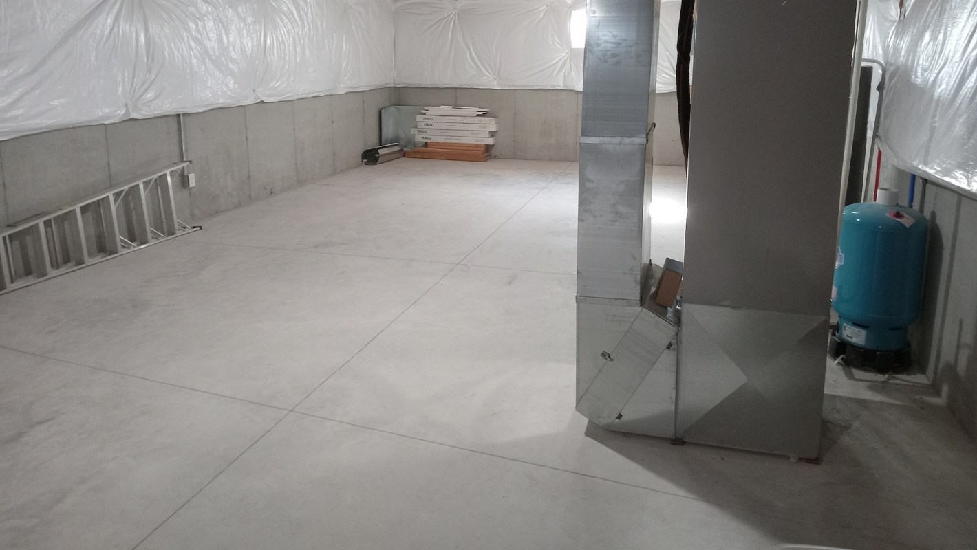 An unfinished basement with concrete floors and walls covered in plastic sheeting, featuring a furnace.