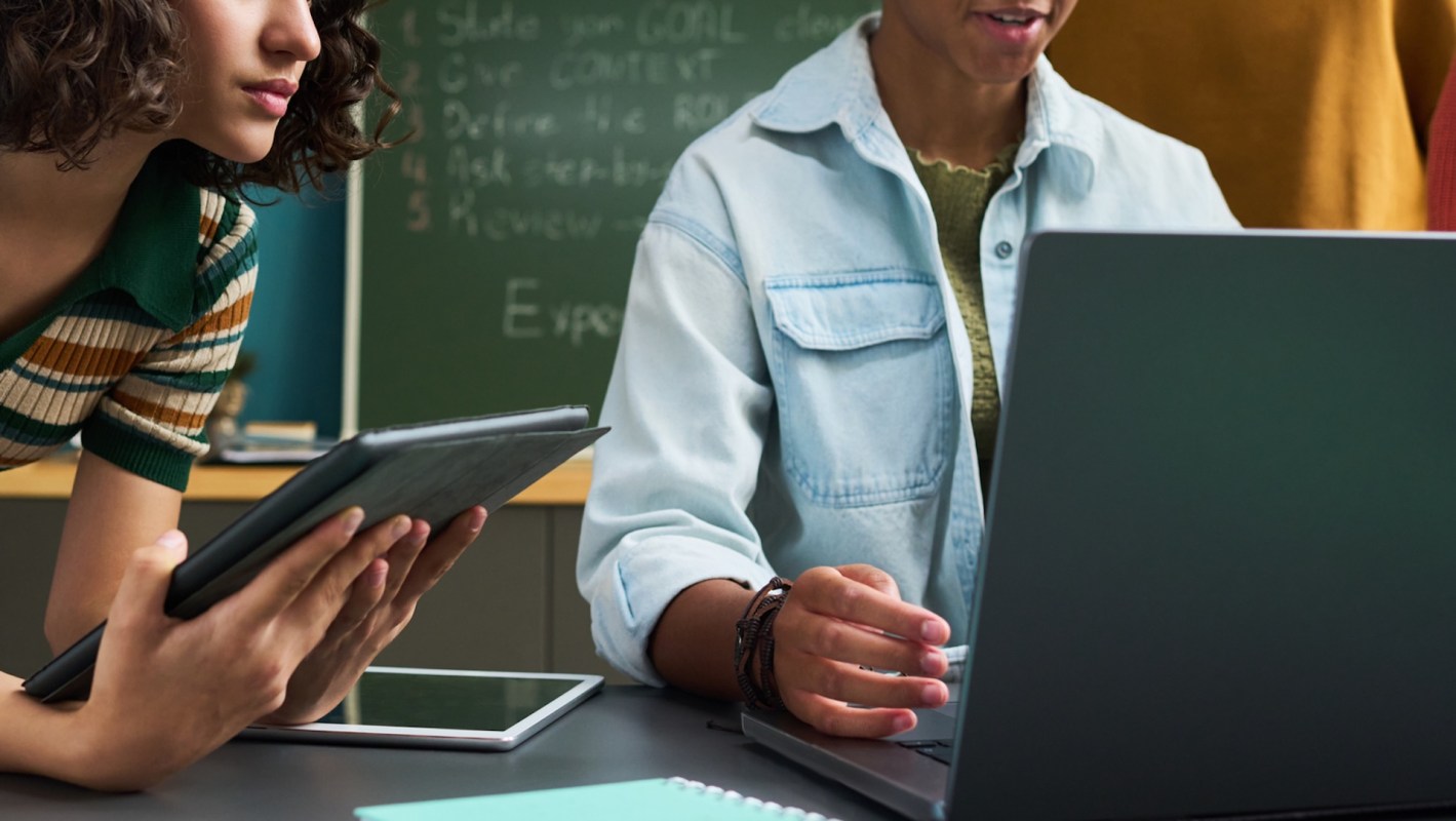 A woman holds a tablet while another person works on a laptop at a table.