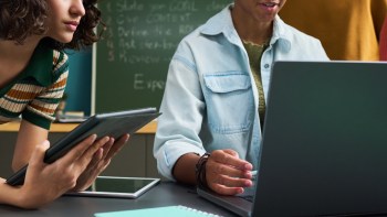 A woman holds a tablet while another person works on a laptop at a table.