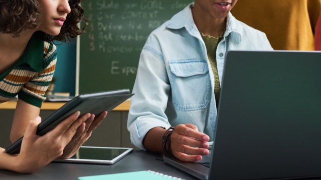 A woman holds a tablet while another person works on a laptop at a table.