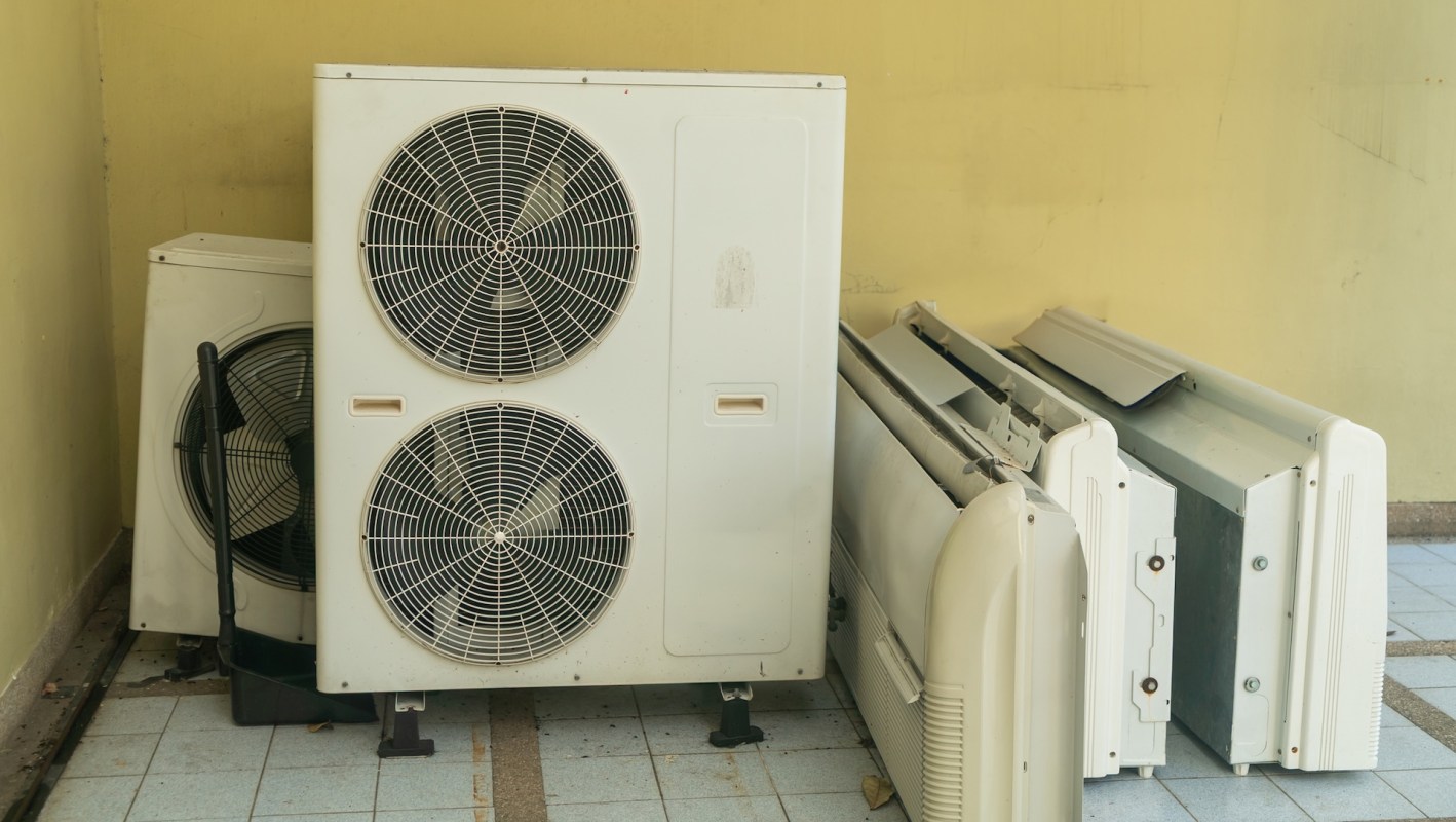 Two air conditioning units and several discarded panels are leaned against a wall with a light-colored background.