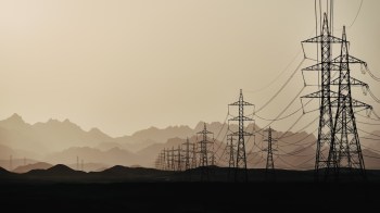 Power lines stretch across a barren landscape with distant mountains under a hazy sky.