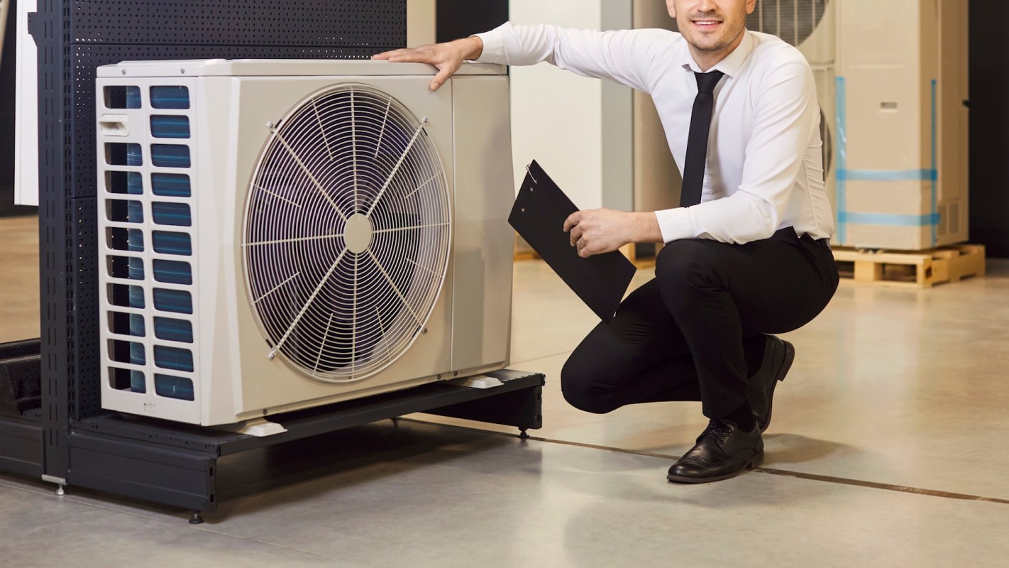 A man crouches beside a large air conditioning unit, holding a clipboard in a warehouse setting.