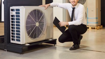 A man crouches beside a large air conditioning unit, holding a clipboard in a warehouse setting.