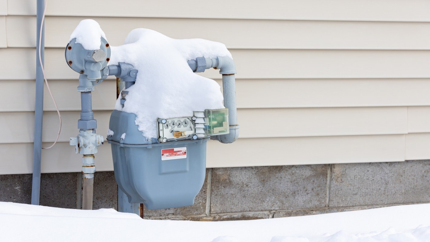 A gas meter covered in snow next to a house's exterior wall.