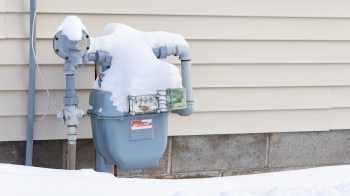 A gas meter covered in snow next to a house's exterior wall.