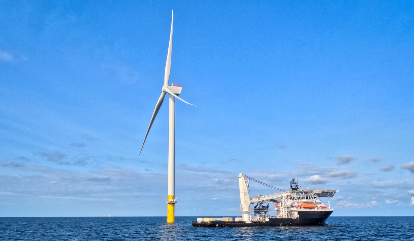 A wind turbine stands next to a service ship in the ocean under a blue sky.