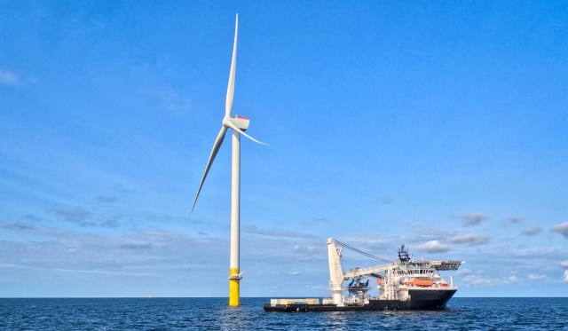 A wind turbine stands next to a service ship in the ocean under a blue sky.