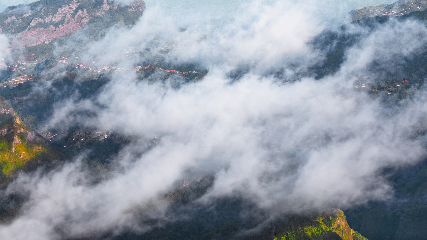 A view of mountains partially obscured by clouds, with hints of greenery and buildings visible below.