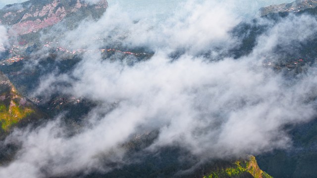 A view of mountains partially obscured by clouds, with hints of greenery and buildings visible below.