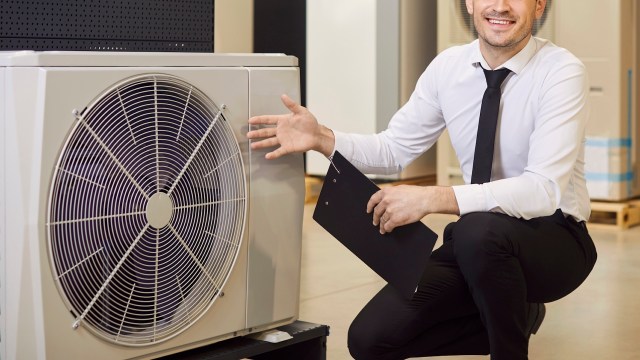 A man poses next to a heat pump unit while holding a clipboard.