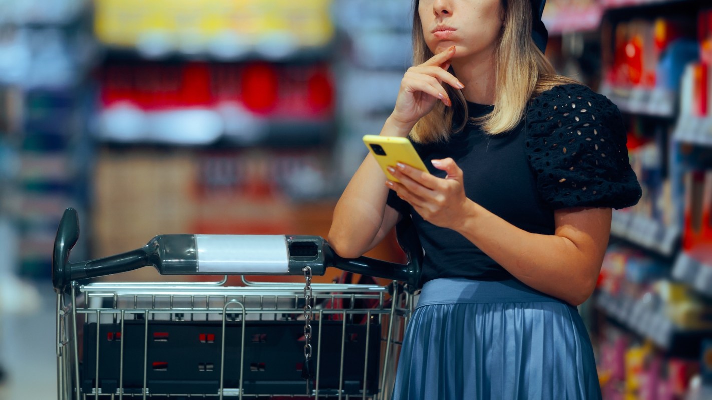 A woman holds a smartphone while standing next to a shopping cart in a grocery store aisle.