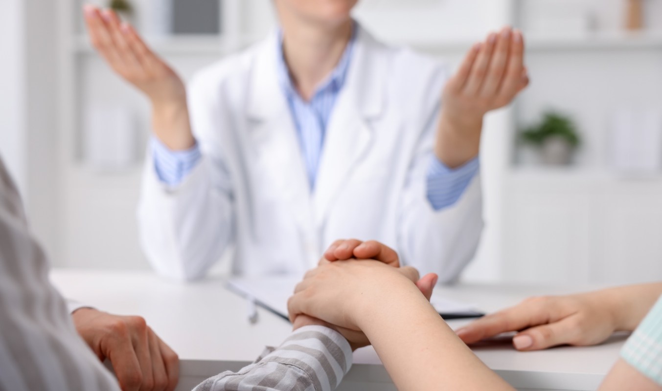 A healthcare professional speaks to two individuals in a consultation setting, hands resting on a table.
