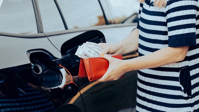 A person holds cash and a red wallet near a car's fuel tank.