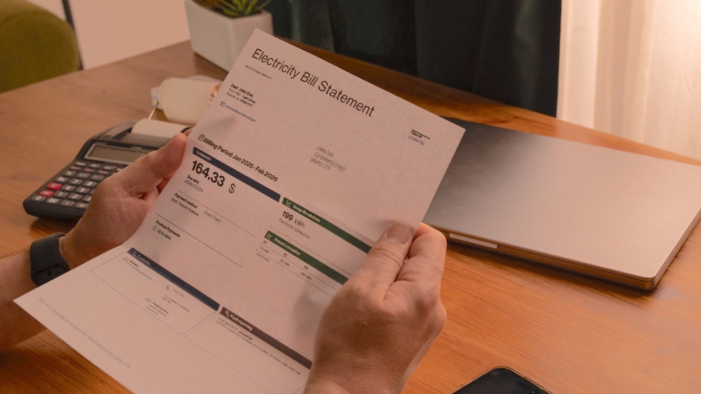 A person holds an electricity bill statement while a calculator and a laptop are on the table.