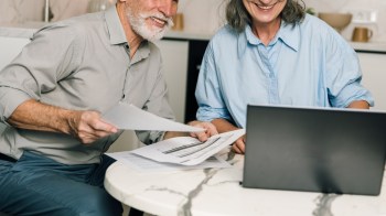 A man and a woman smile at a table with papers and a laptop, engaged in discussion.