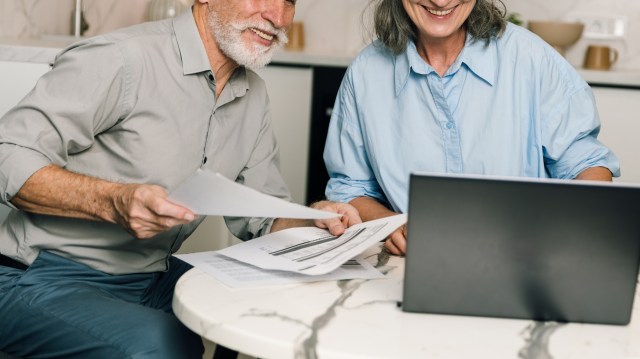 A man and a woman smile at a table with papers and a laptop, engaged in discussion.