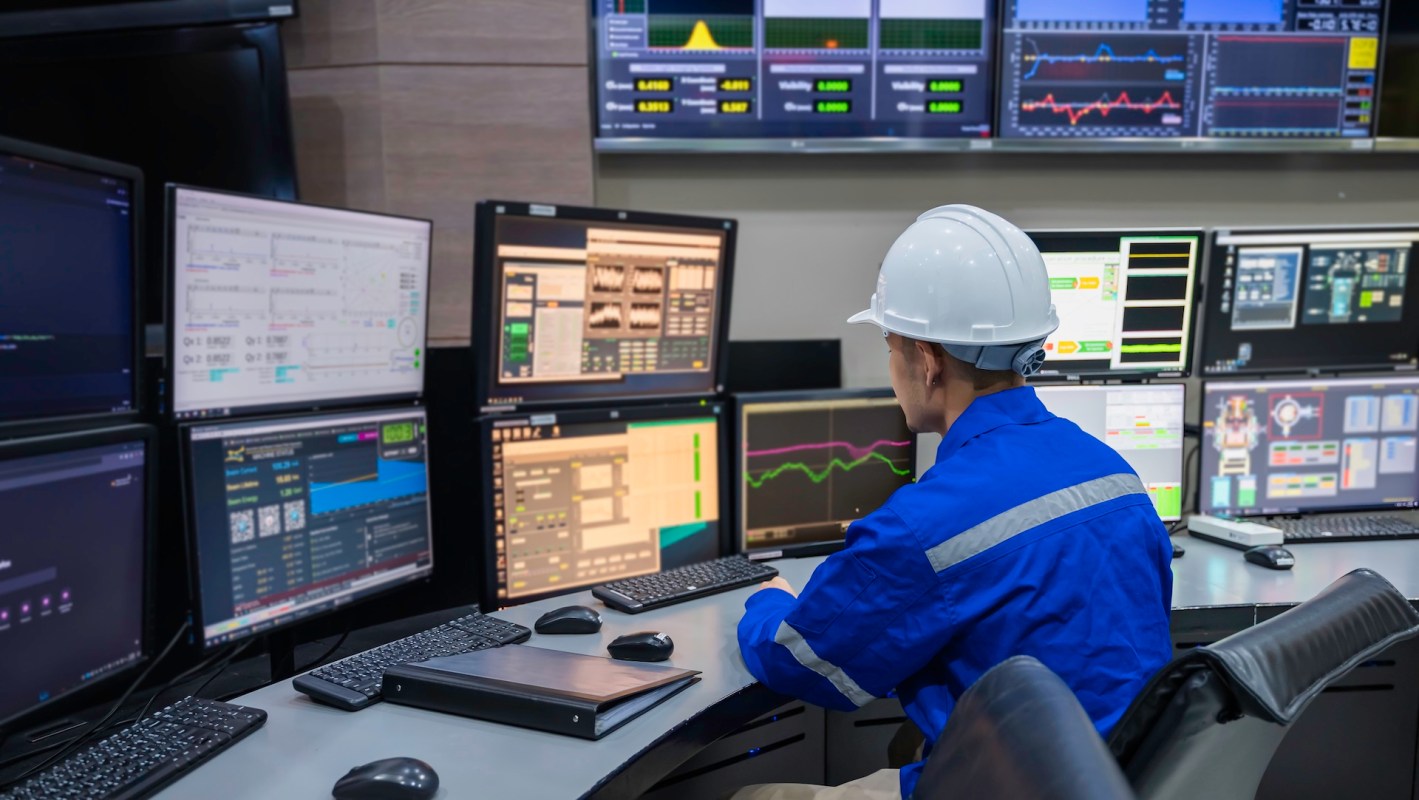 A worker in a blue uniform and helmet monitors multiple screens in a control room.
