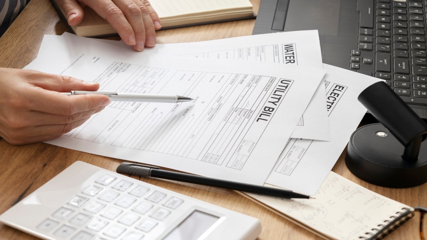 A person's hand holds a pen over utility bills on a wooden desk with a calculator and a laptop nearby.
