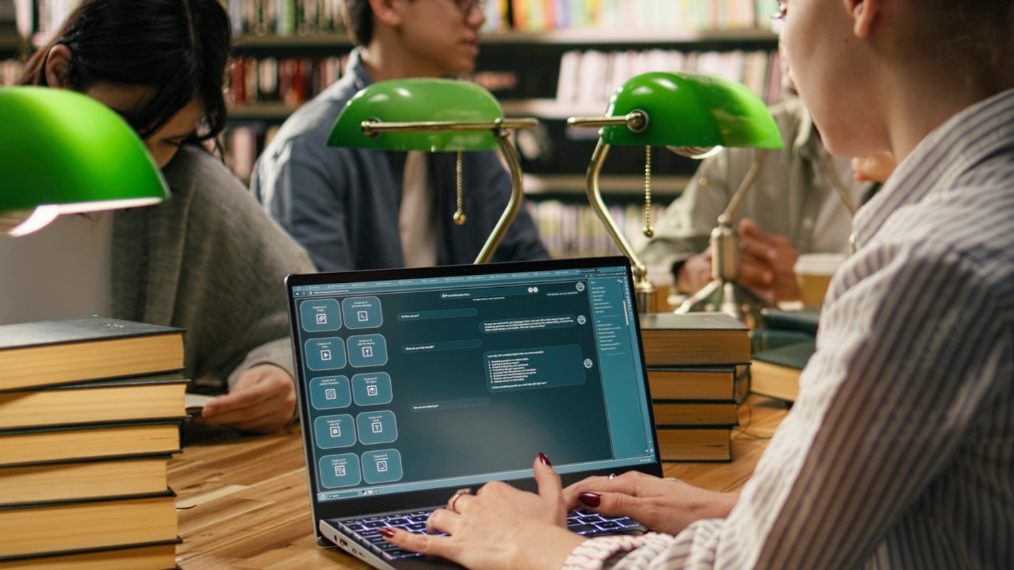 A laptop displaying a digital interface is placed on a wooden table surrounded by people in a library.