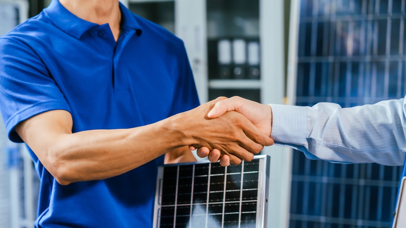 Two individuals shake hands next to a solar panel.