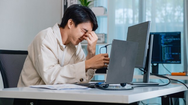 A person sitting at a desk, looking frustrated while working on a laptop with two monitors in the background.