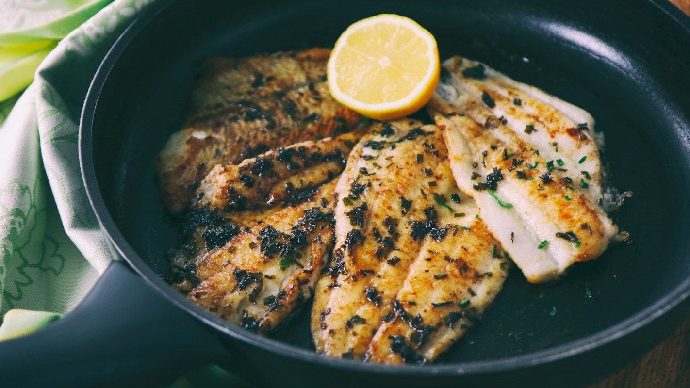 Cooked flounder fillets garnished with herbs and a lemon slice in a frying pan.