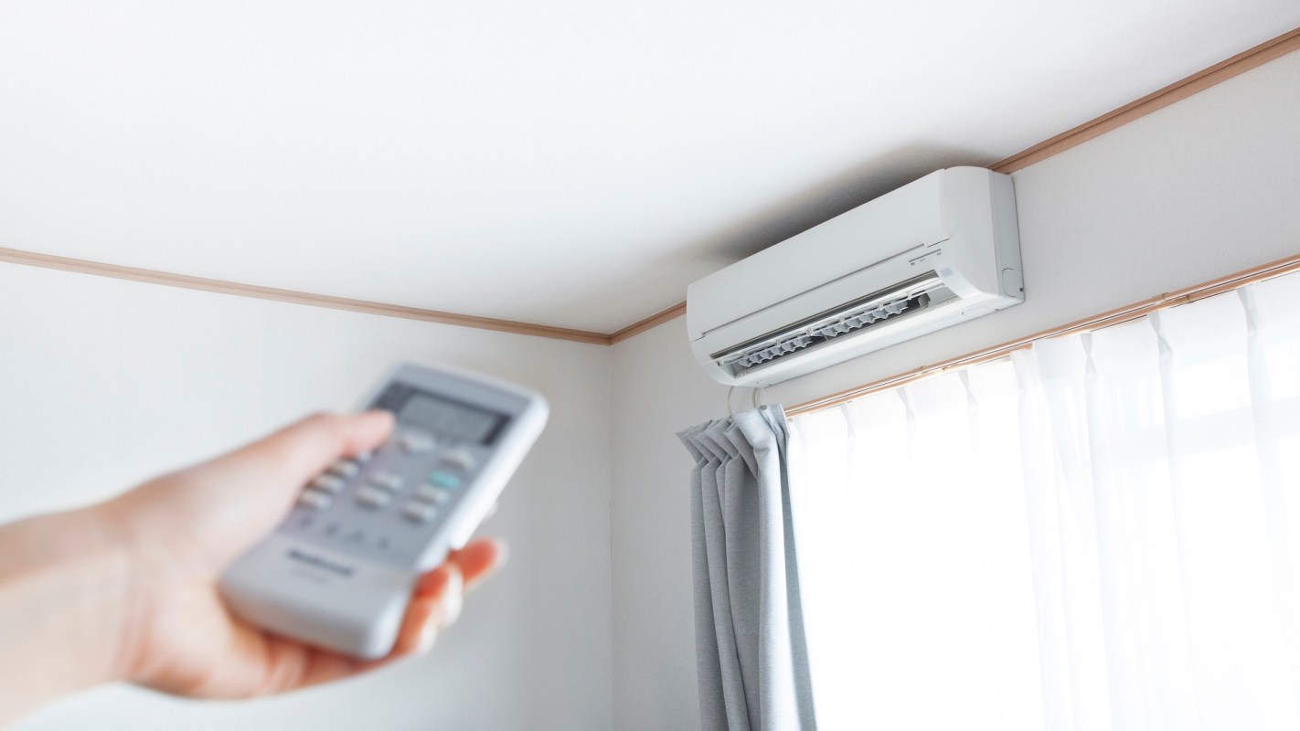 A hand holding a remote control points towards a wall-mounted air conditioner.