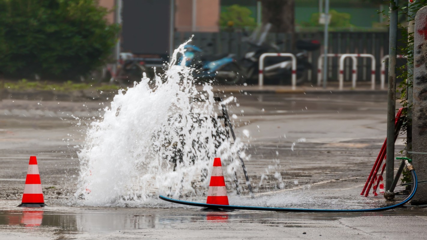 A burst water main splashes water onto a street near traffic cones and a hose.