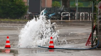 A burst water main splashes water onto a street near traffic cones and a hose.