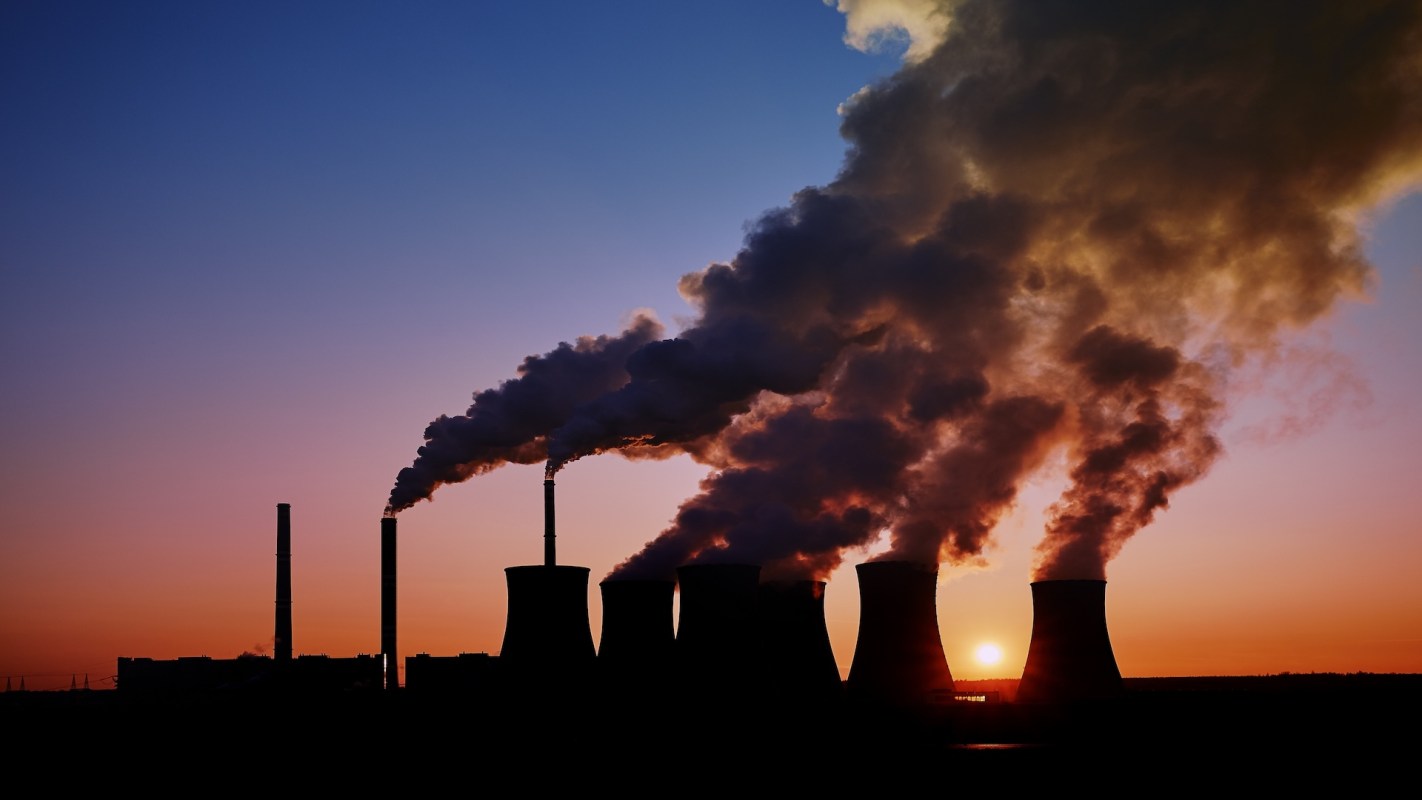 A power plant with cooling towers emitting smoke against a sunset sky.