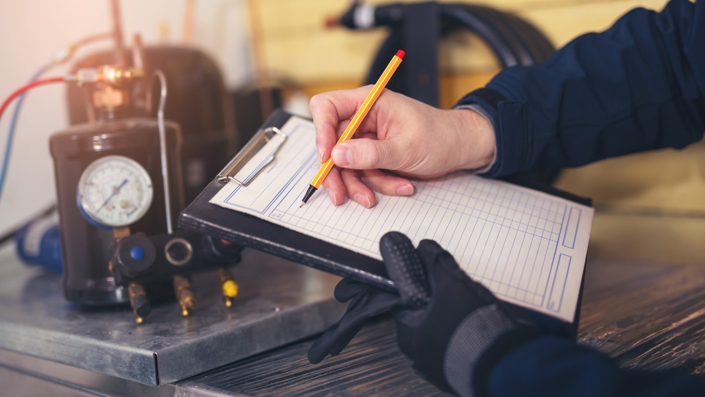 A hand holding a pen writes on a clipboard next to a pressure gauge and a compressor.