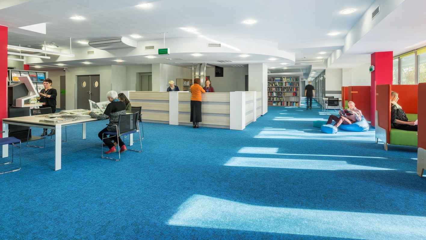 A modern library interior with a blue carpet, seating areas, and people engaging in various activities.