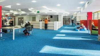 A modern library interior with a blue carpet, seating areas, and people engaging in various activities.